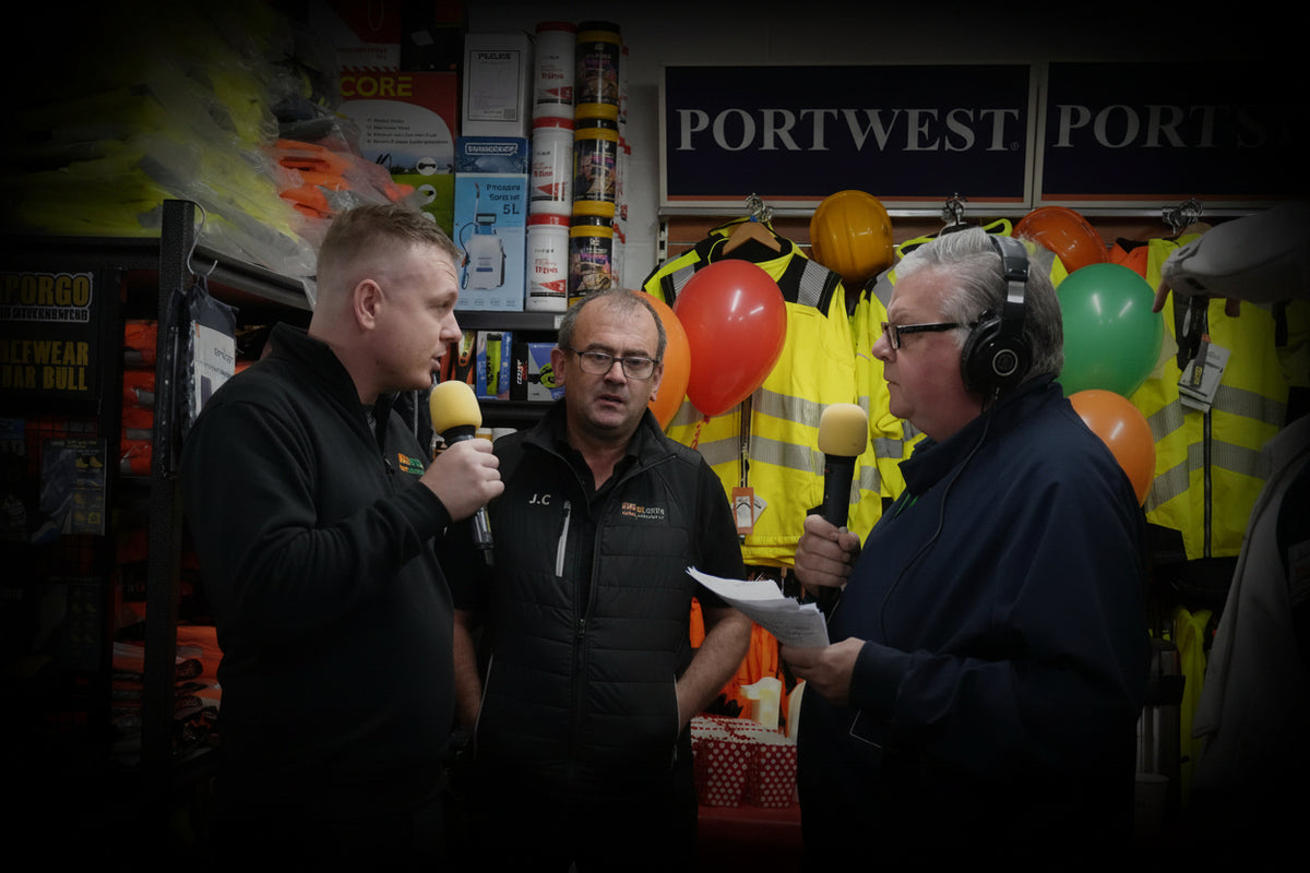 Three men in a retail store holding microphones, with high-visibility jackets, balloons, and Portwest signage in the background.